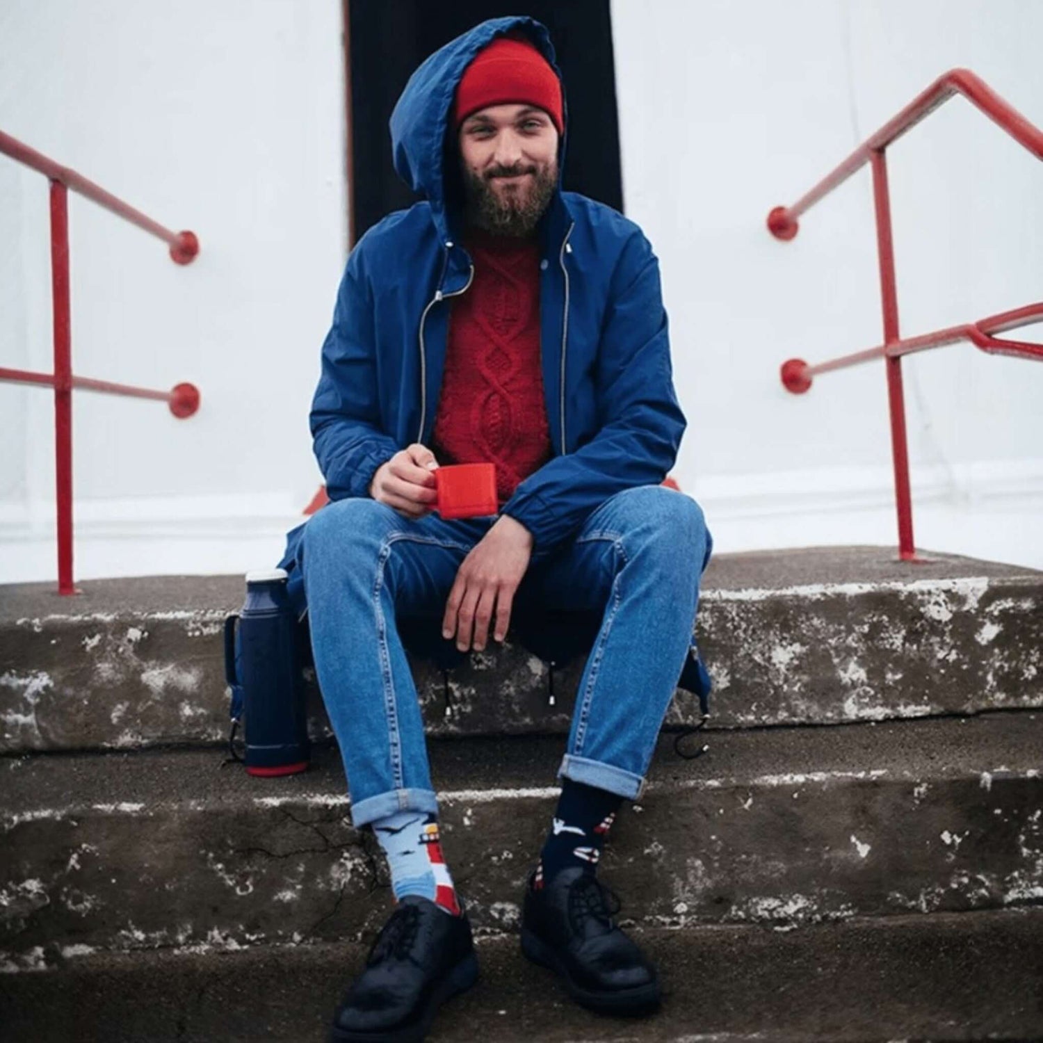 Homme assis sur des marches portant des chaussettes fantaisie dépareillées à motifs phares et mouettes, look marin avec bonnet rouge et veste bleue.