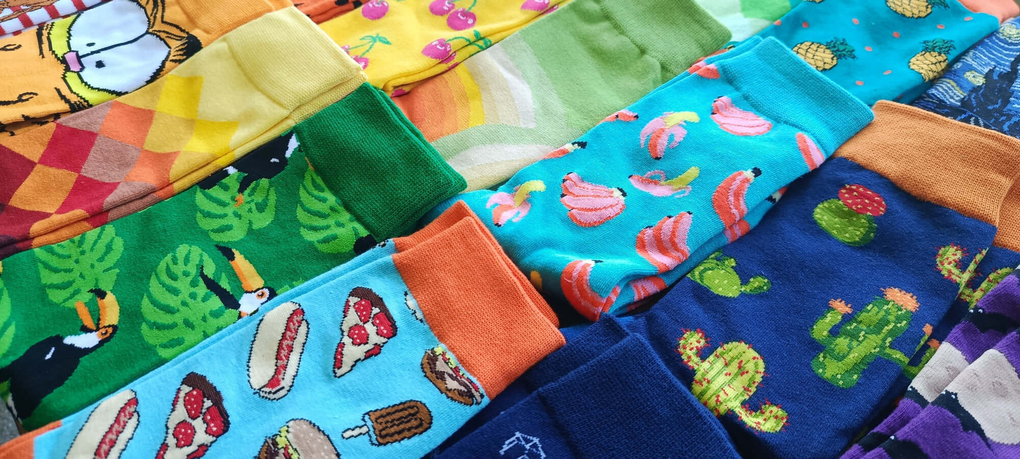 Collection colorée de chaussettes fantaisie avec des imprimés tropicaux, des fruits et des designs ludiques, présentée dans une mise en scène vibrante.